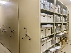Photo of archive storage area; on the left, the hand cranks to operate shelving units; on the right, the shelves of one unit containing storage boxes