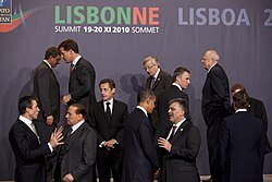 Twelve men in black suits stand talking in small groups under a backdrop with the words Lisbonne and Lisboa.