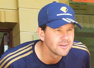 A man stands in front of a brick wall; he is wearing a cap.