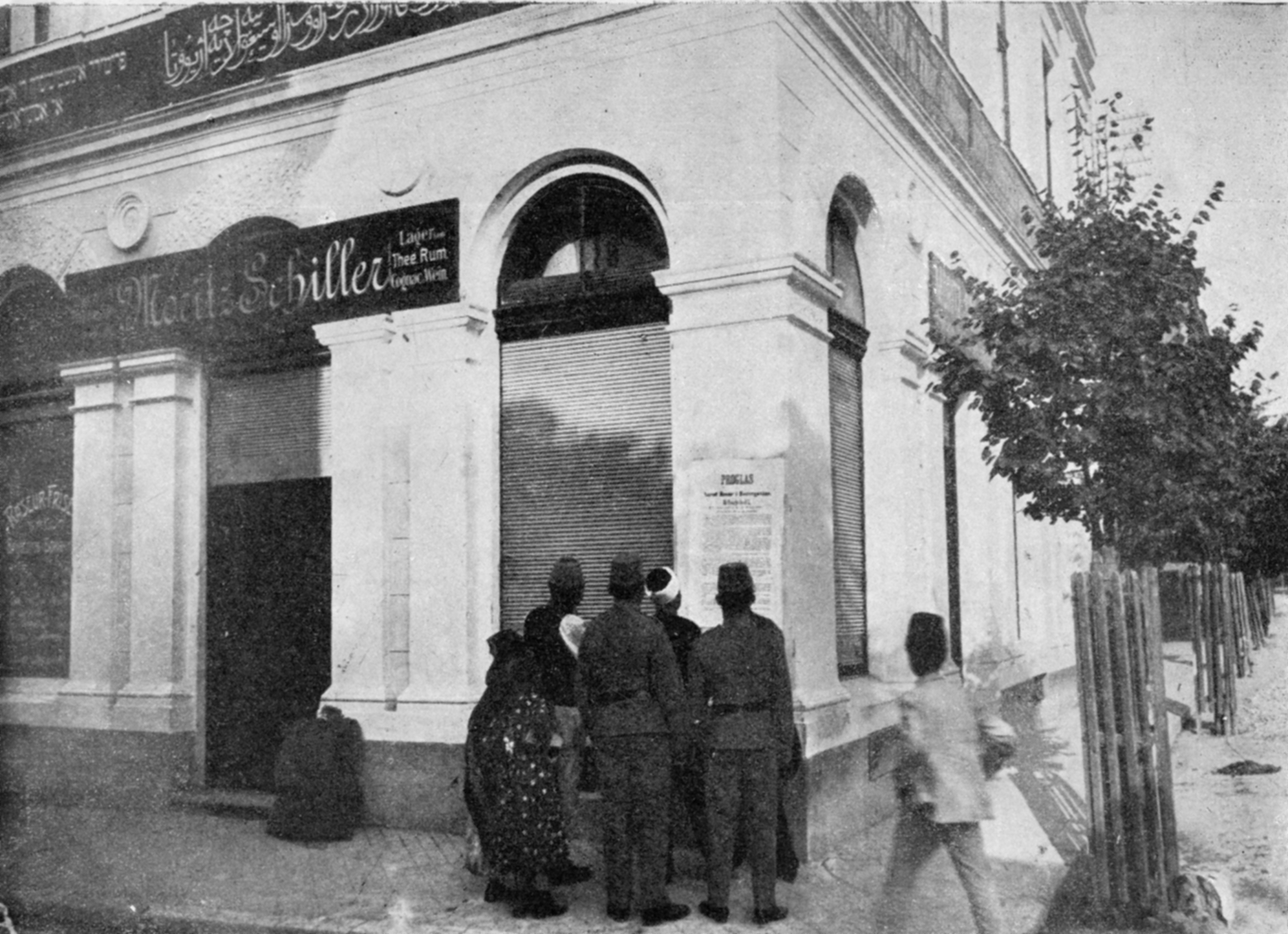 Photo of large white building with one signs saying "Moritz Schiller" and another in Arabic; in front is a cluster of people looking at poster on the wall.
